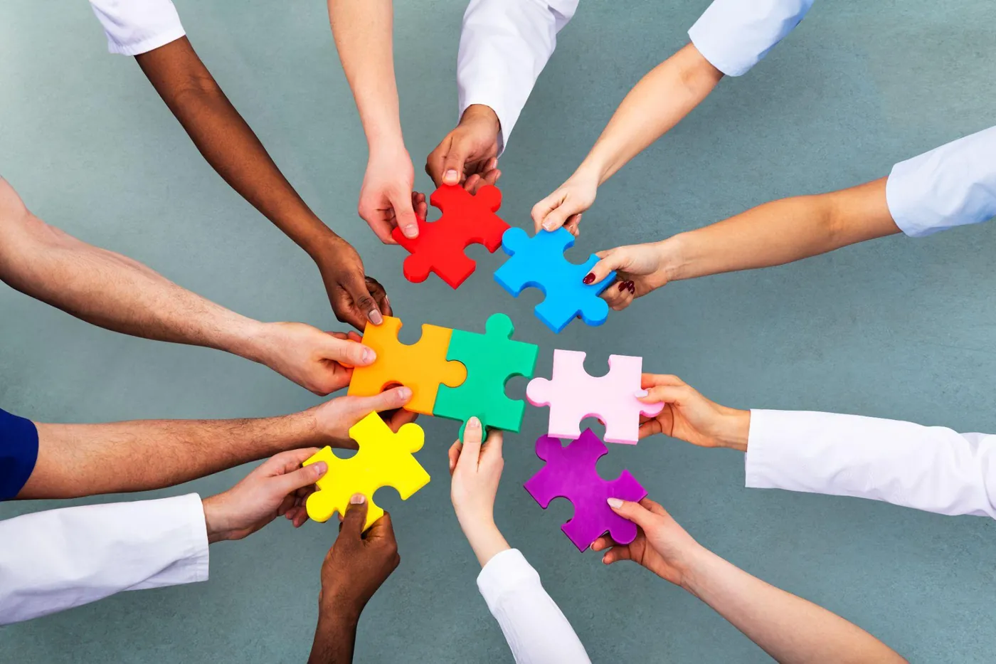 Many hands of different skin tones joining colorful puzzle pieces together.
